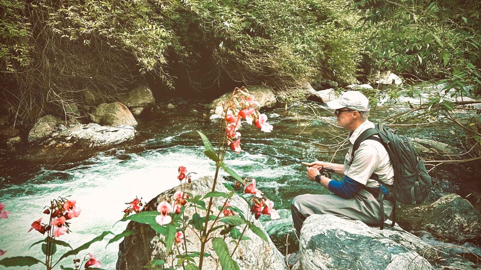 An angler taking a break, sitting on a rock beside a clear alpine stream in the Dolomites, with pink wildflowers in the foreground.